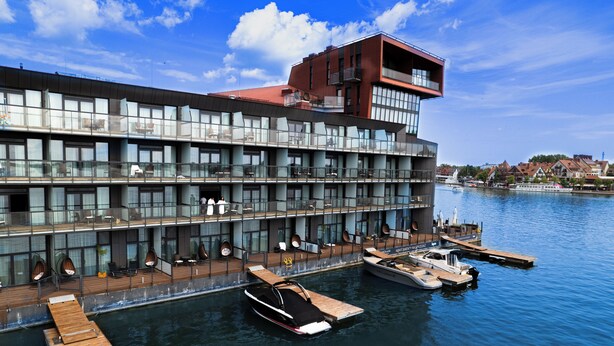 A modern hotel with multiple balconies overlooking a body of water, a dock with boats, and a clear view of the surrounding town. - Hotel Mikołajki Leisure & SPA - Destigo Hotels (Mikolajki)