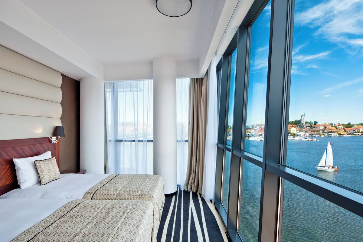Hotelkamer met een groot raam, een bed, nachtkastje en uitzicht op een waterlichaam met een zeilboot.