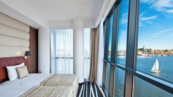 Hotelkamer met een groot raam, een bed, nachtkastje en uitzicht op een waterlichaam met een zeilboot.