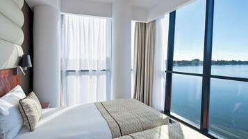 A modern hotel room with a large bed, a nightstand, a lamp, and a view of a lake through large windows.