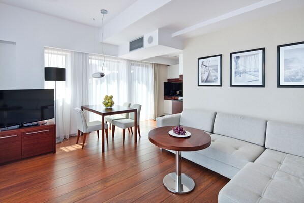 Una sala moderna con televisión de pantalla plana, suelo de madera, una mesa de comedor con sillas, una mesa redonda de centro con fruta, un sofá gris y cuadros en la pared.