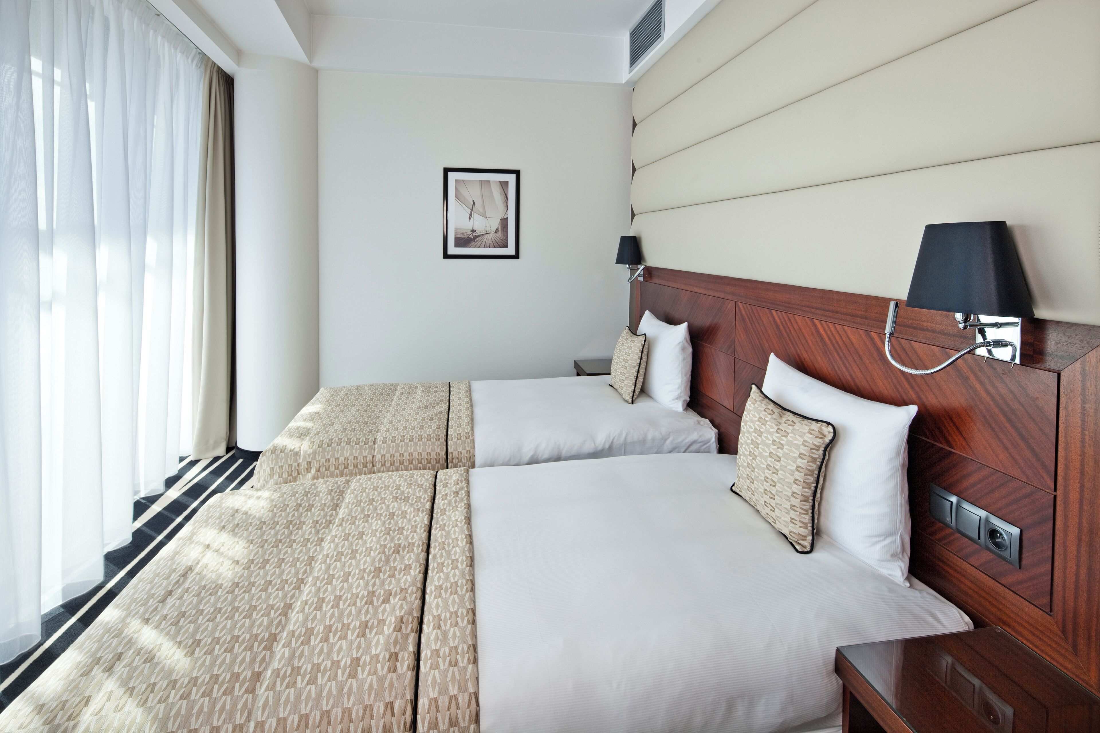 Habitación de hotel con dos camas, cabecero de madera, mesita de noche, lámpara y un cuadro enmarcado en la pared.