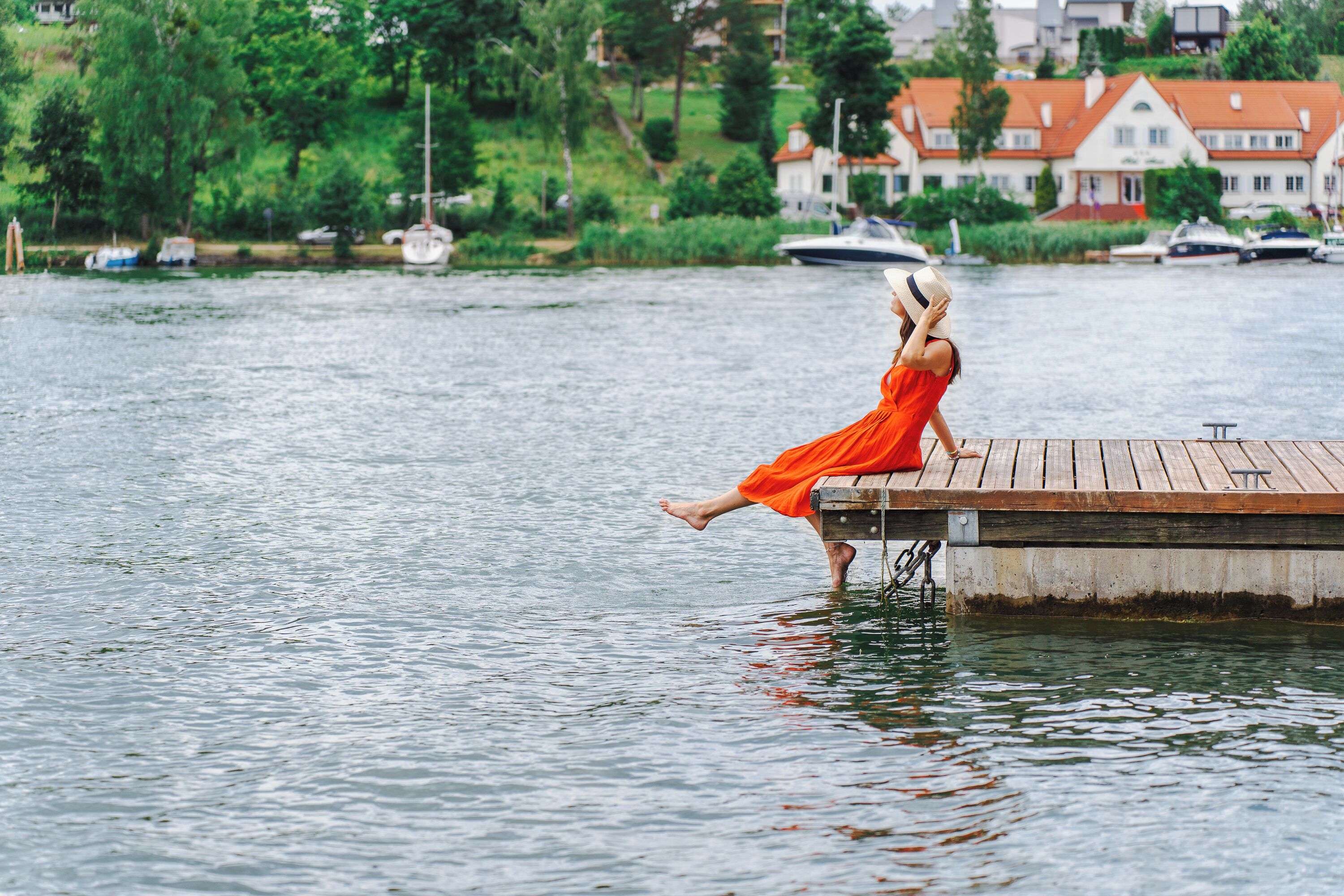 A woman in an orange dress sitting on a wooden dock by a calm lake with boats and houses in the background.