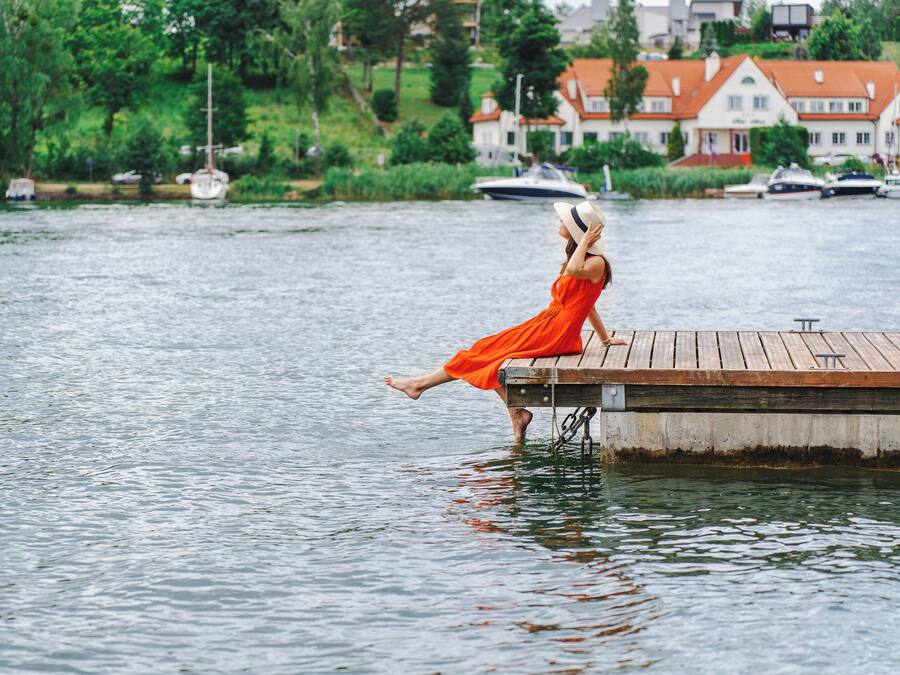 A woman in an orange dress sitting on a wooden dock by a calm lake with boats and houses in the background.