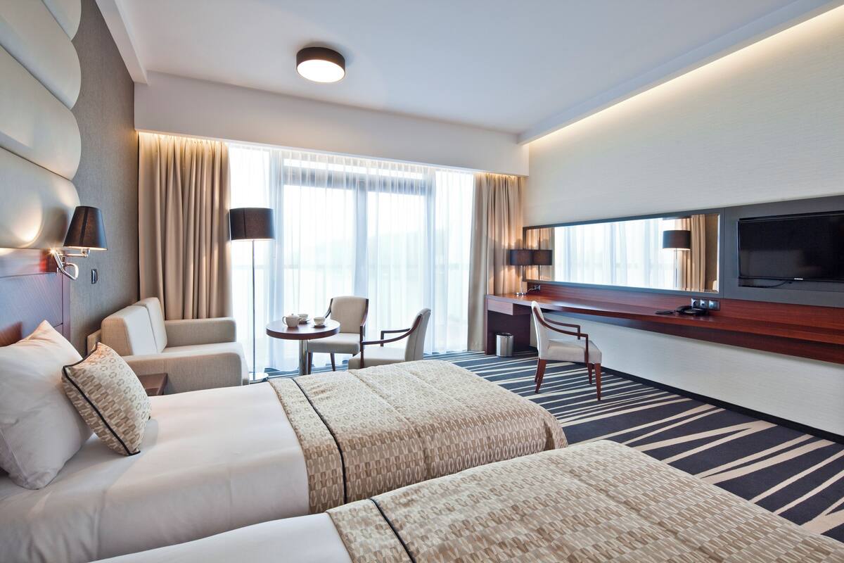 Hotelkamer met twee bedden, een bureau, een stoel, een televisie en een groot raam met gordijnen.
