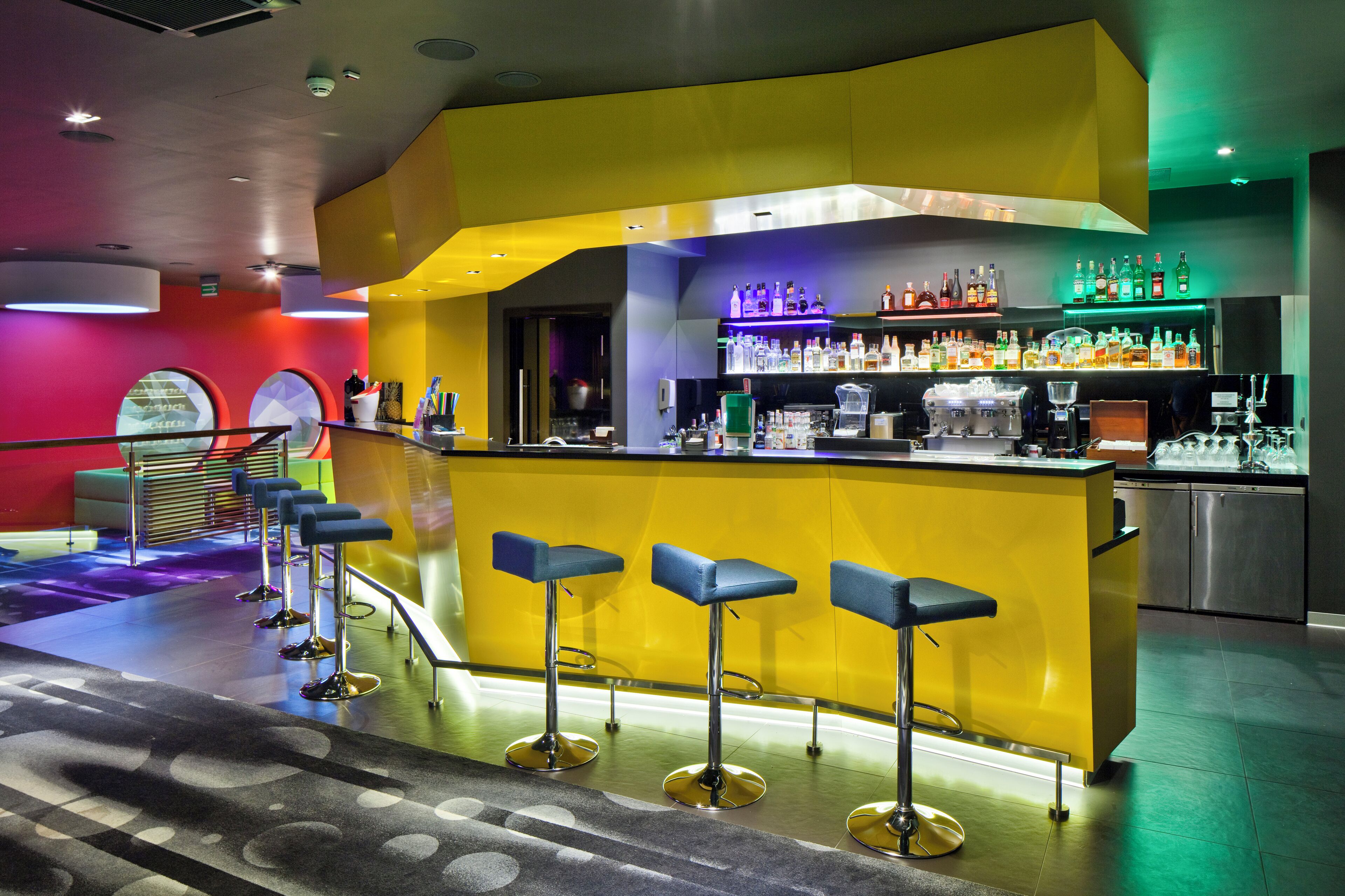 Una zona de bar con un mostrador amarillo, varios taburetes y una gran variedad de botellas y vasos.