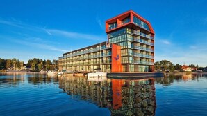A modern hotel with a distinctive red and orange facade, large windows, and a reflection in the water. - Hotel Mikołajki Leisure & SPA - Destigo Hotels (Mikolajki)