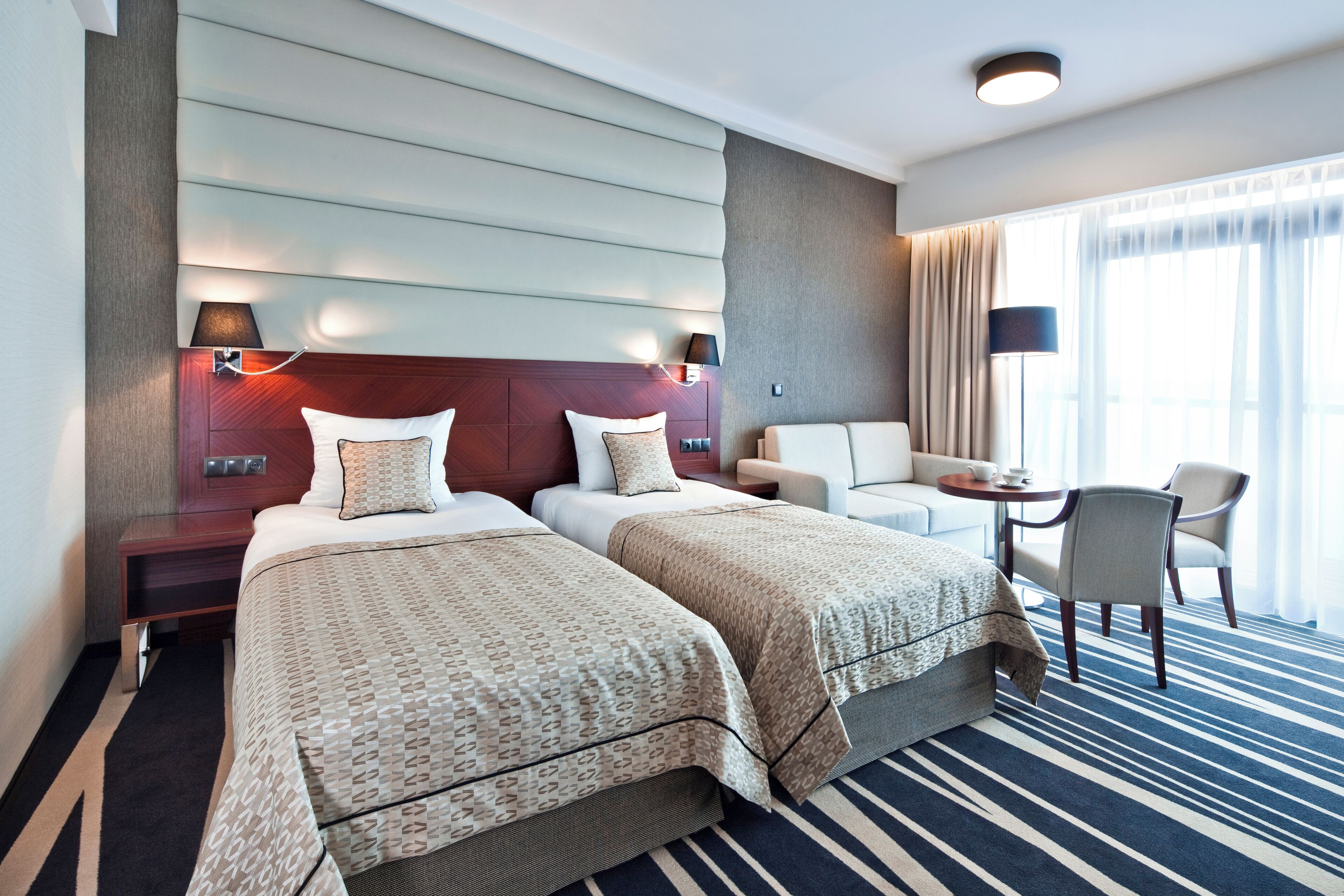 A hotel room with two beds, a sofa, a small table, and a view of the outdoors.
