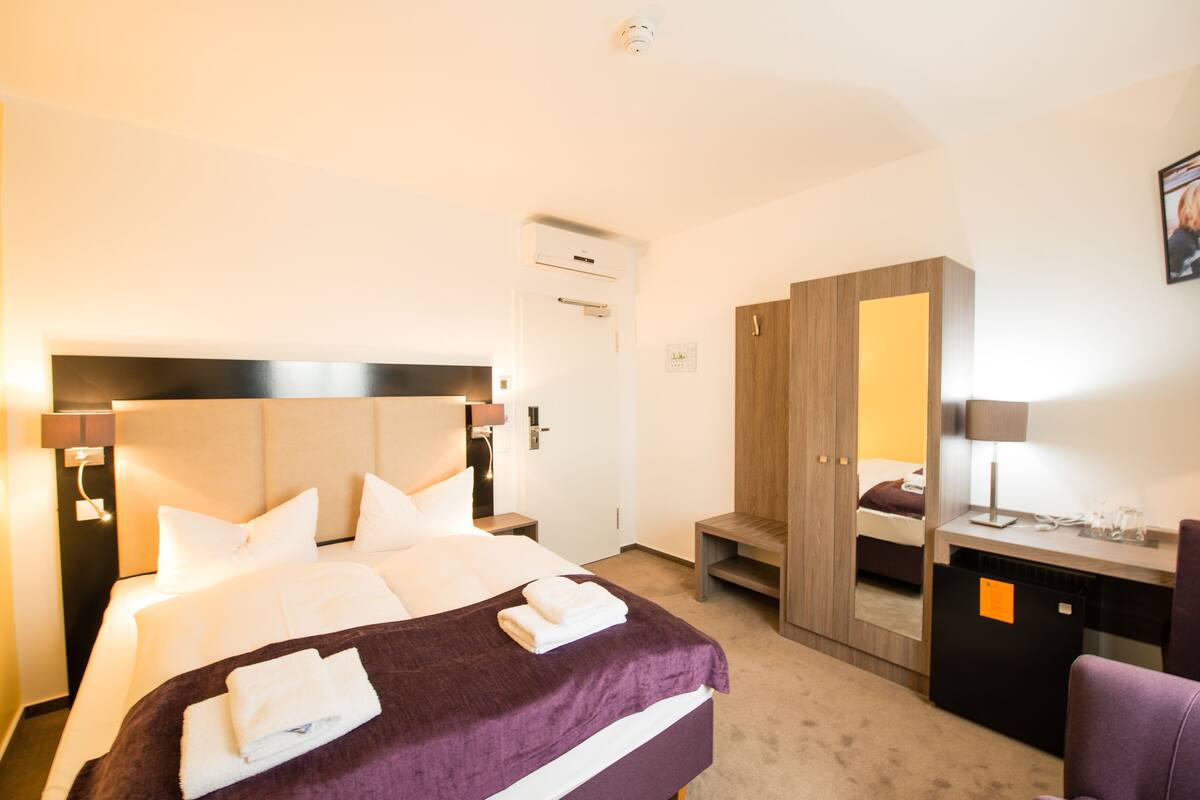 A hotel room with a large bed, a desk with a chair, a wardrobe, and a mirror.