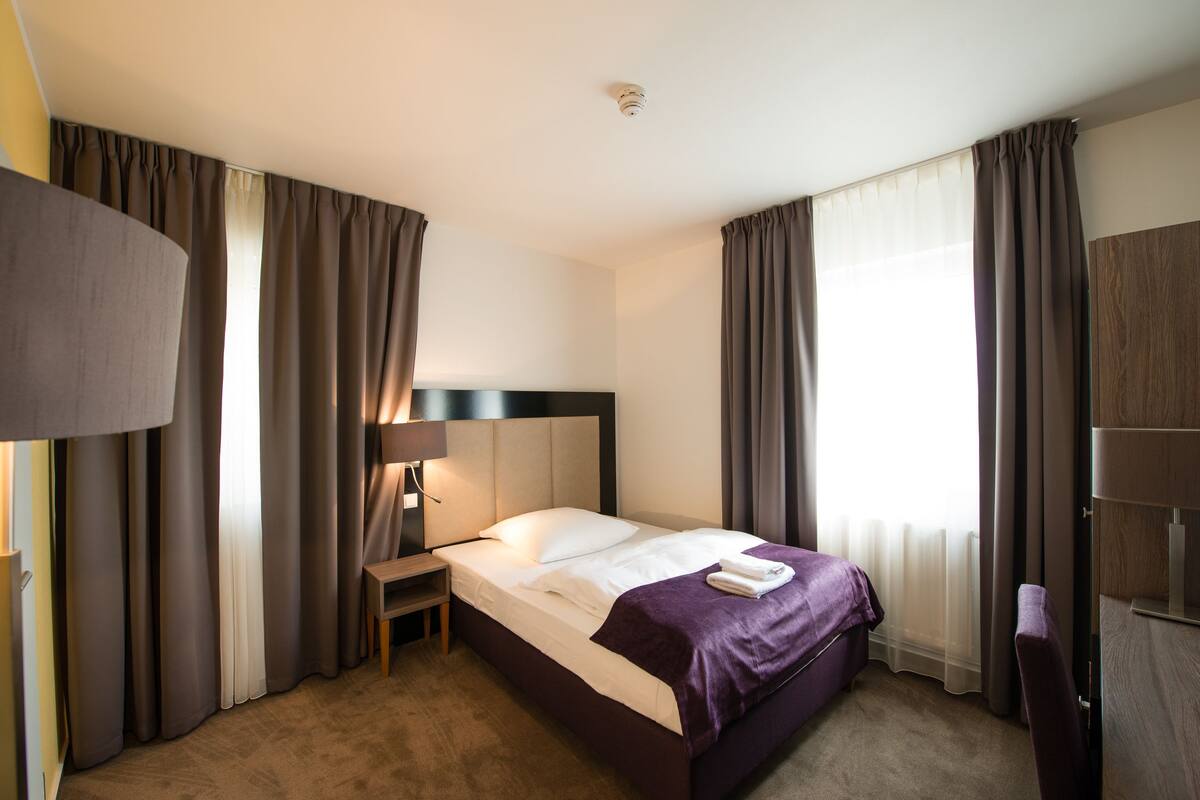 A hotel room with a bed, a nightstand, a lamp, and a window with curtains.