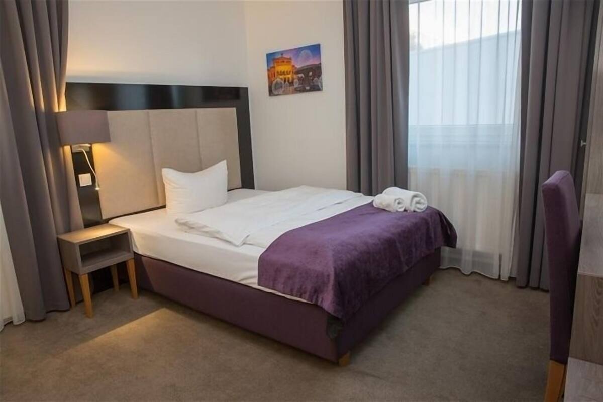 A hotel room with a bed, a nightstand, a chair, a painting on the wall, and a window with curtains.