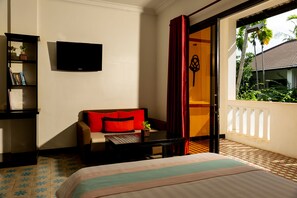 A hotel room with a bed, a sofa, a TV, a bookshelf, and a balcony with a view of palm trees. - La Residence WatBo Hotel (Siem Reap)