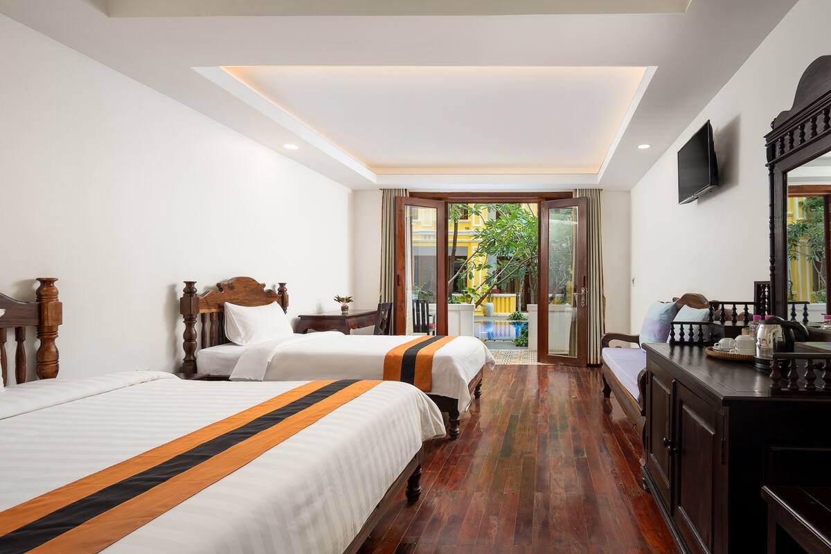 A hotel room with two beds, a wooden floor, a desk with a chair, a TV, and a balcony with a view of greenery.