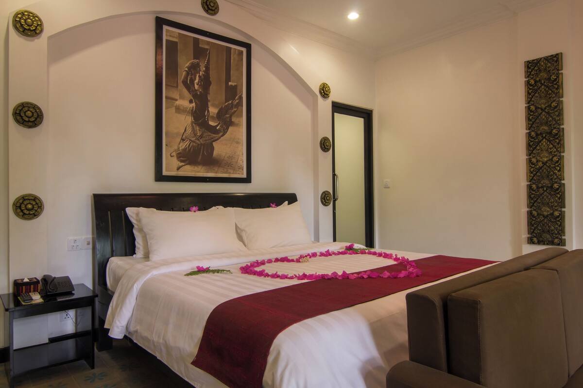A neatly made bed with a red blanket and pink flower petals, a framed picture above it, and a brown armchair to the right.