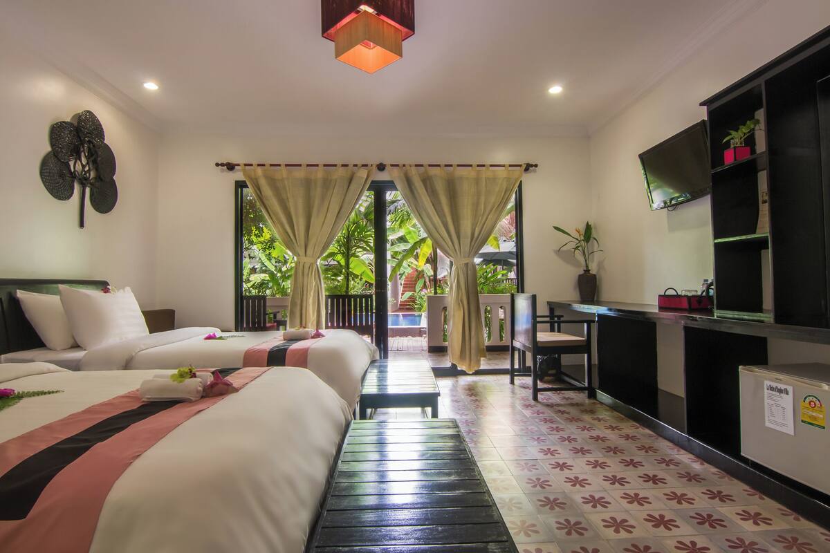 A hotel room with two beds, a desk, a TV, and a view of greenery outside.