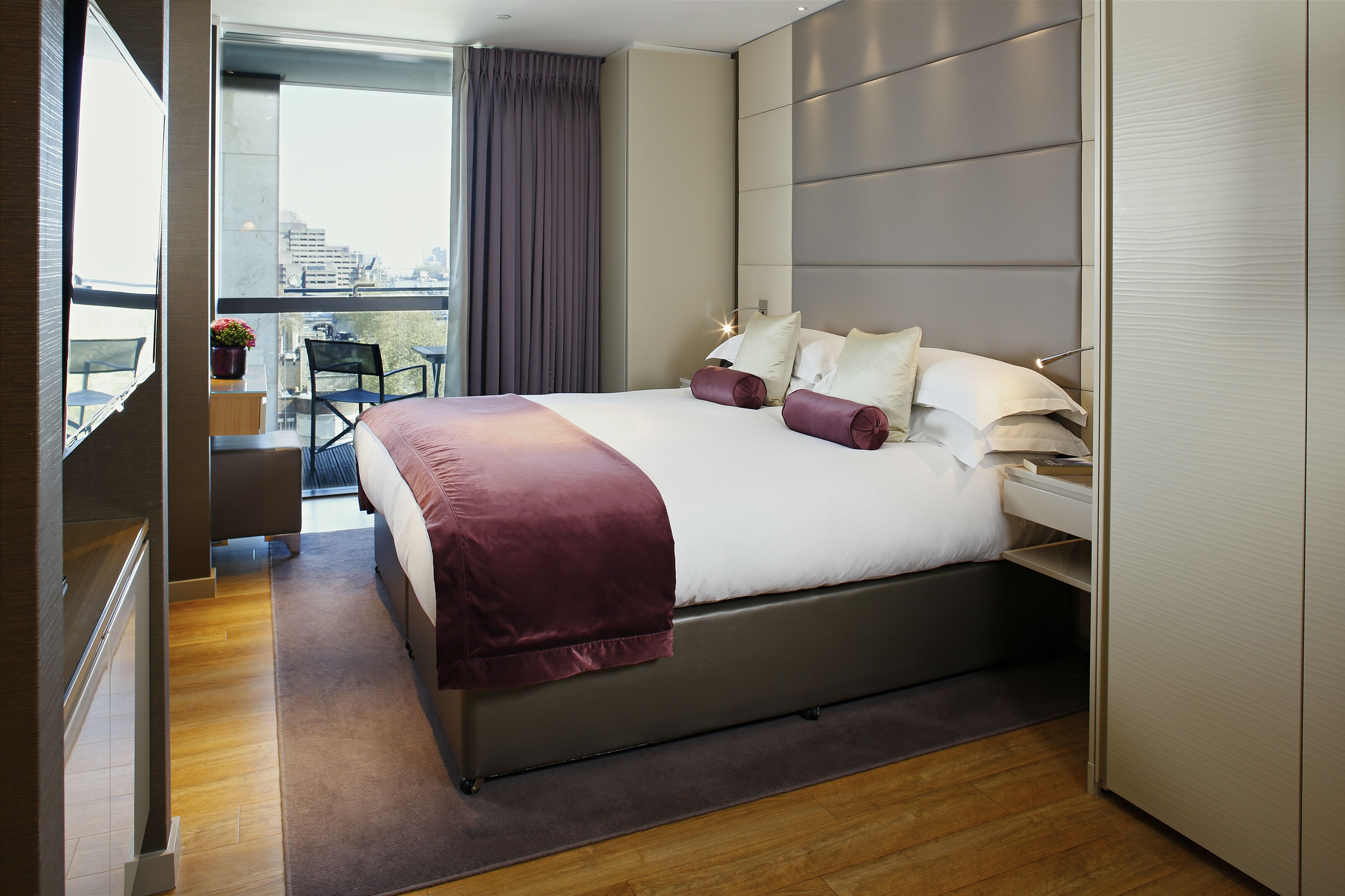A modern bedroom with a large bed, a view of the city, and a balcony with a chair and potted plant.