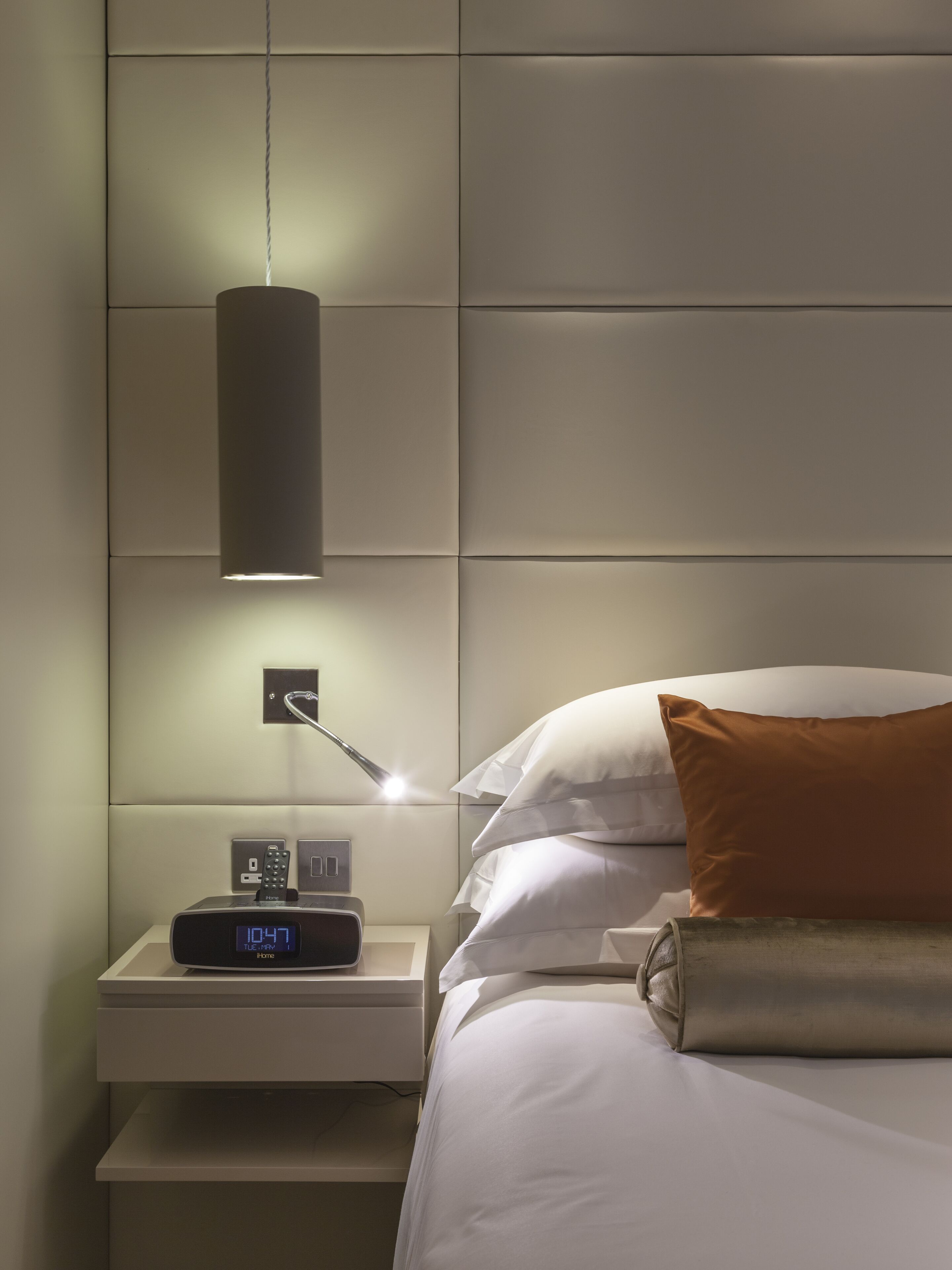 A modern hotel room with a bed, a bedside table with a digital clock, a lamp, and a power outlet.