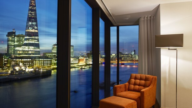 A modern room with a large window offering a view of a city skyline at dusk, a plush orange armchair, and a floor lamp. - Cheval Three Quays at The Tower of London (London)
