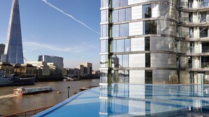 A modern building with a curved facade and large windows, overlooking a river with a boat and a tall skyscraper in the background. - Cheval Three Quays at The Tower of London (London)