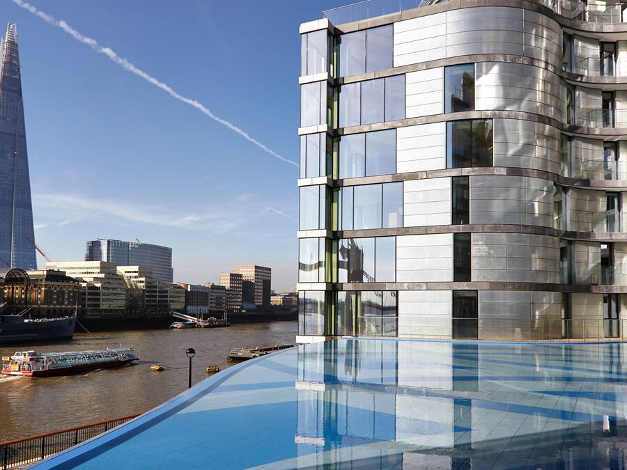 A modern building with a curved facade and large windows, overlooking a river with a boat and a tall skyscraper in the background.