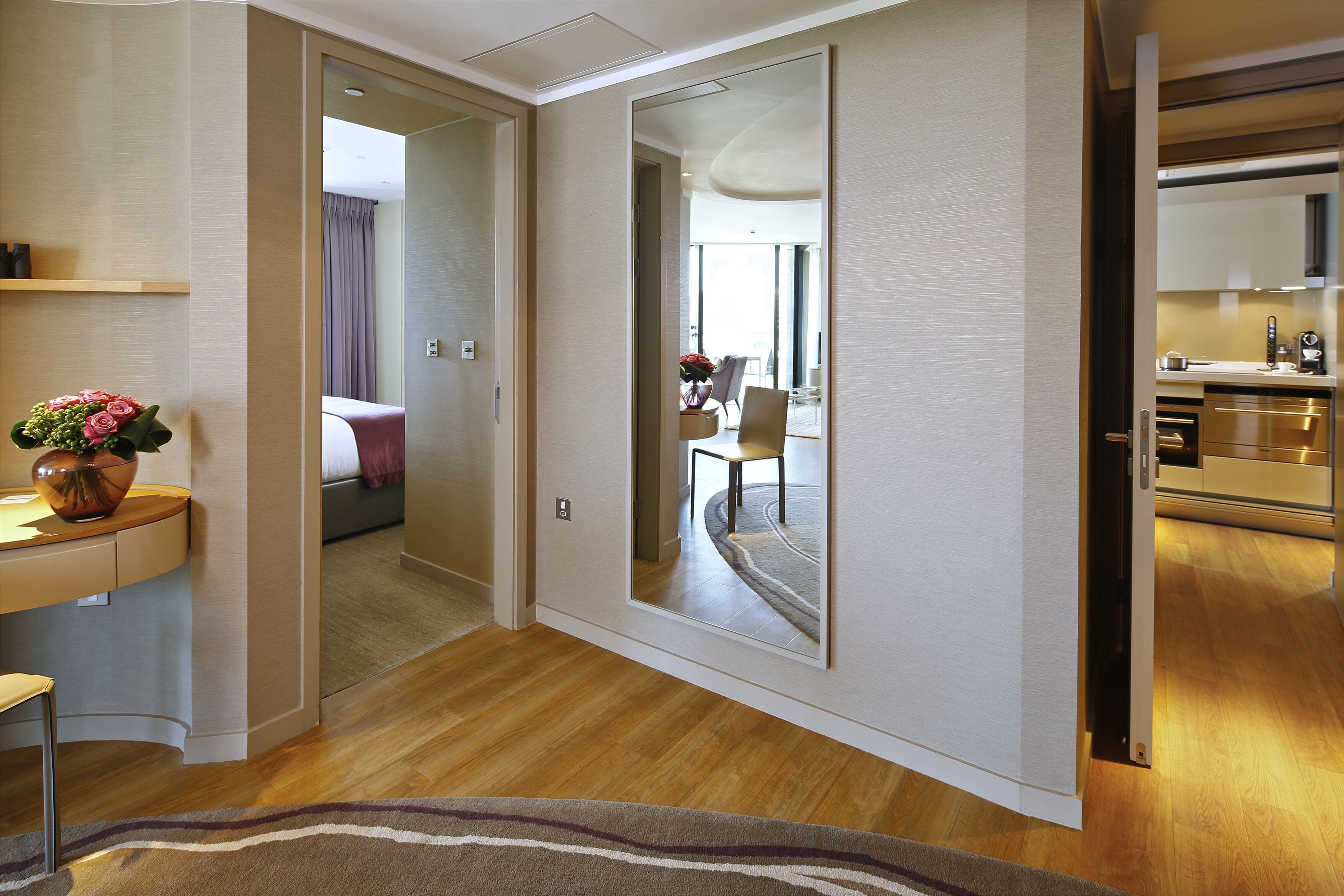 Quarto de hotel moderno com piso de madeira, um espelho grande, uma escrivaninha com um vaso de flores e vista para a área de refeições através de uma porta aberta.