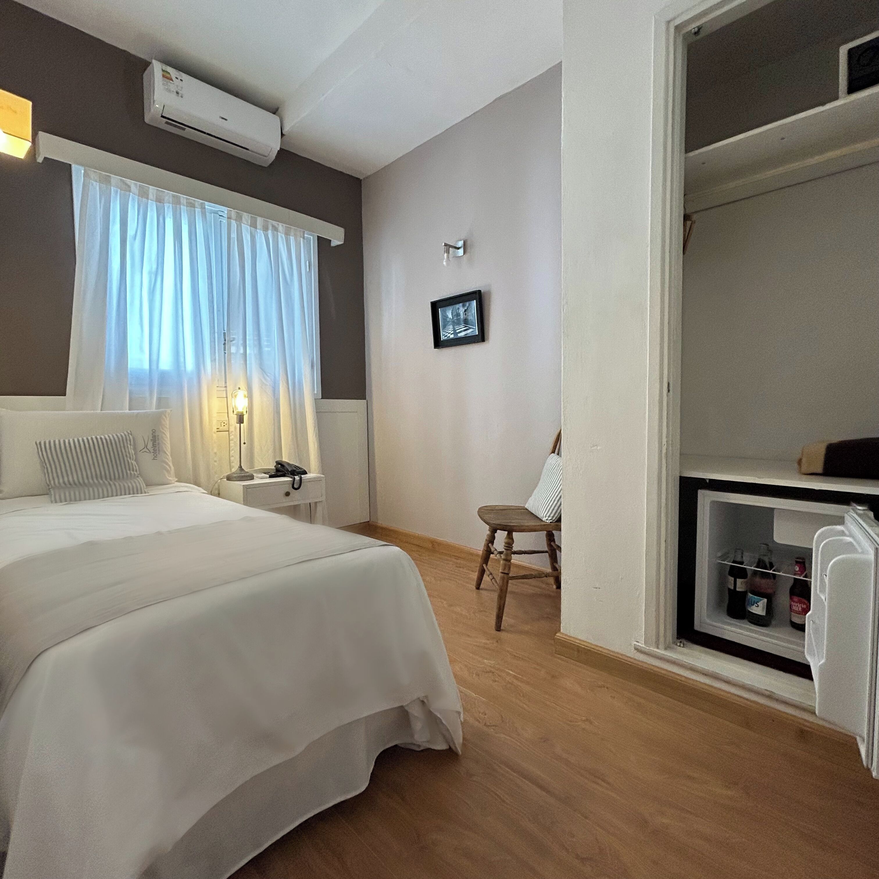 Una habitación de hotel con cama, mesita de noche, lámpara, silla y un pequeño estante con botellas.