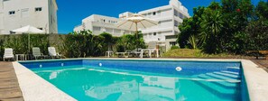 Una piscina rectangular con borde de azulejos, rodeada por un deck de madera. Se observan sillones de descanso y una mesa con sombrilla al fondo.