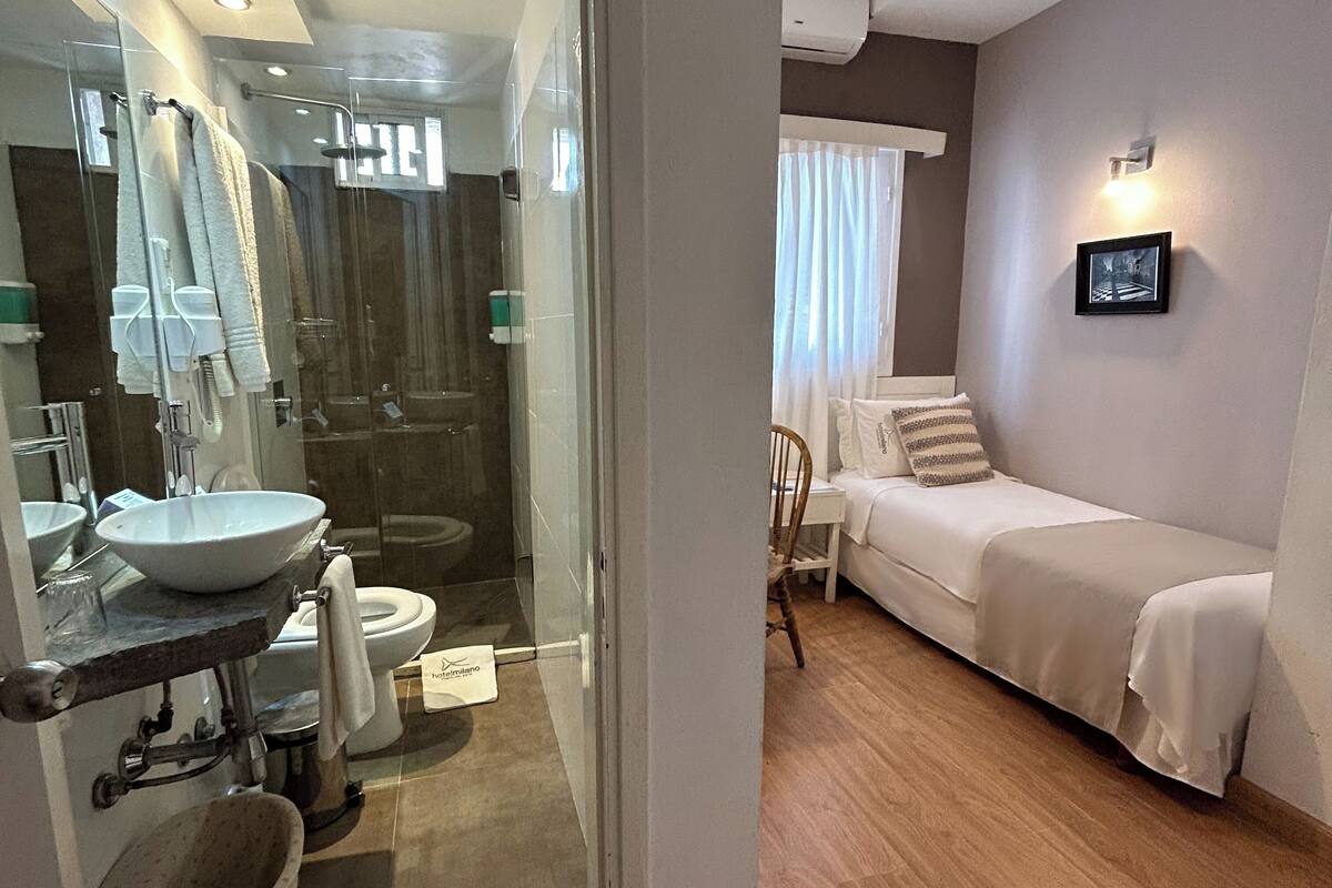 A hotel bathroom with a sink, toilet, and shower. There is a bed with a pillow and a wall-mounted picture.