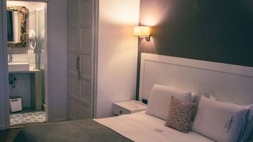 Habitación de hotel con cama, mesitas de noche, un espejo, una lámpara colgante y un baño visible a través de una puerta abierta.