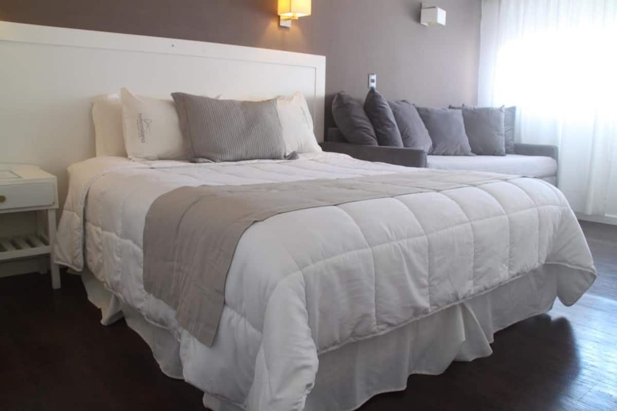 A neatly made bed with a white and gray duvet, a bedside table with a lamp, and a dark wooden floor.