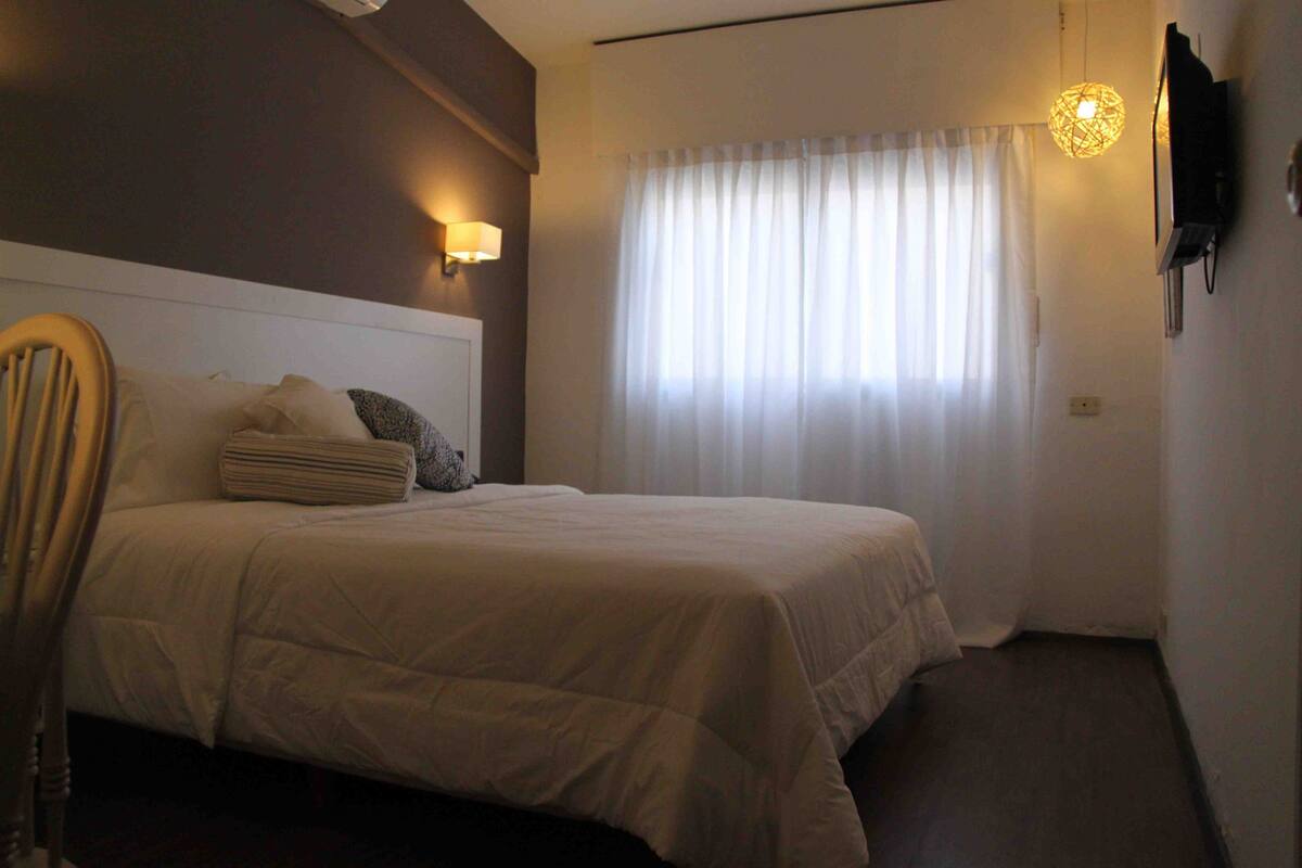 A hotel room with a bed, a chair, a lamp, and a window with curtains.