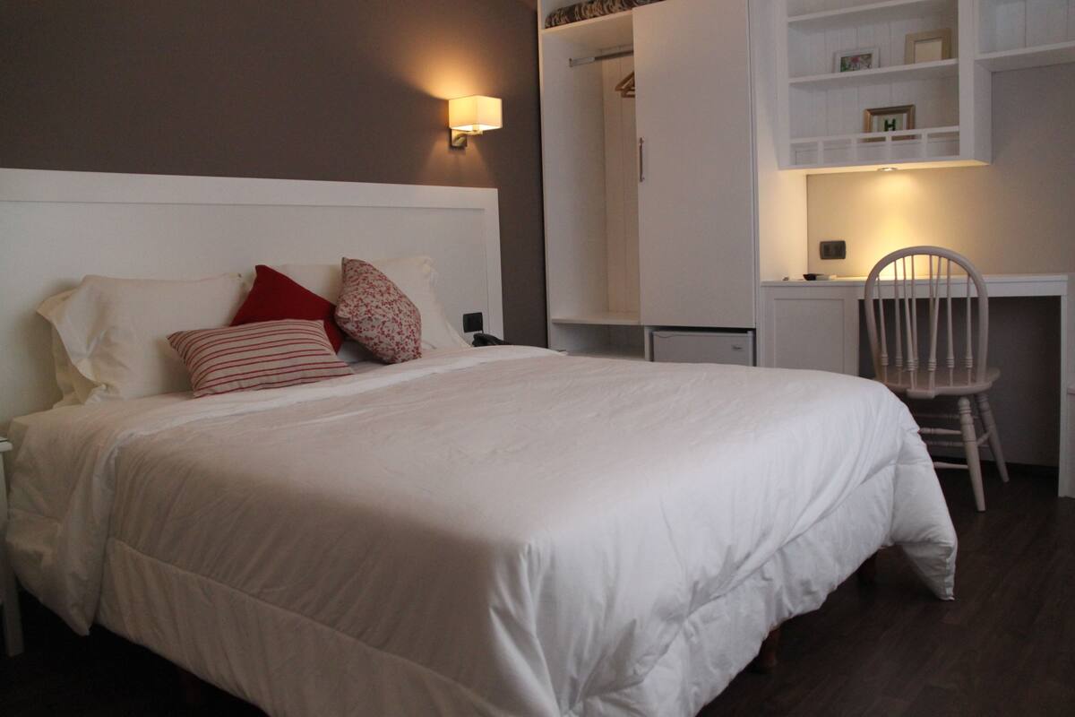 A neatly made bed with white bedding and red pillows, a wooden chair, and a white wardrobe with a mirror and shelves.