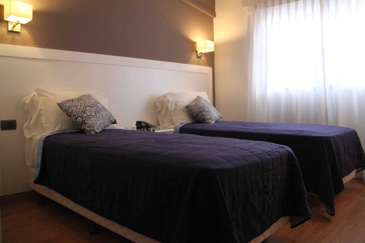 A hotel room with two beds, each with a dark-colored bedspread and pillows, a white headboard, and wall-mounted lamps.
