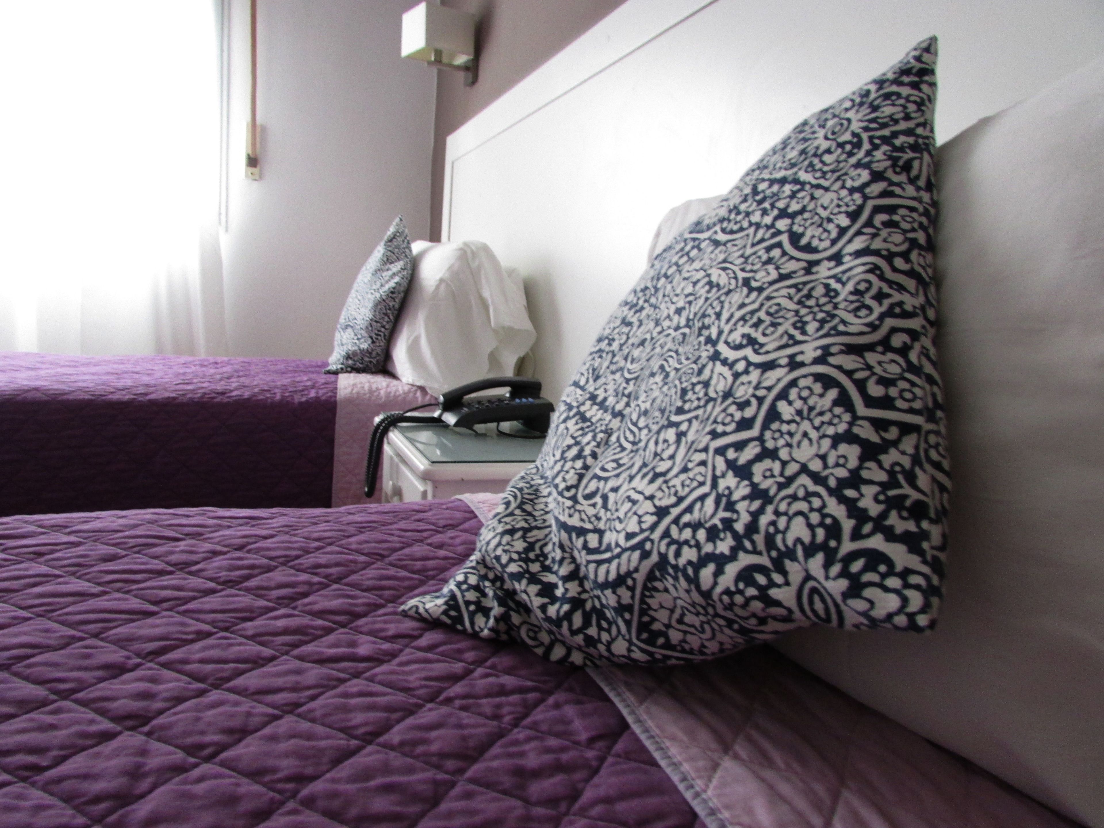 Habitación de hotel con dos camas, cada una con una colcha morada acolchada. Hay un almohadón estampado en una cama y un almohadón blanco en la otra. Un teléfono se ve sobre una mesita de noche junto a las camas.
