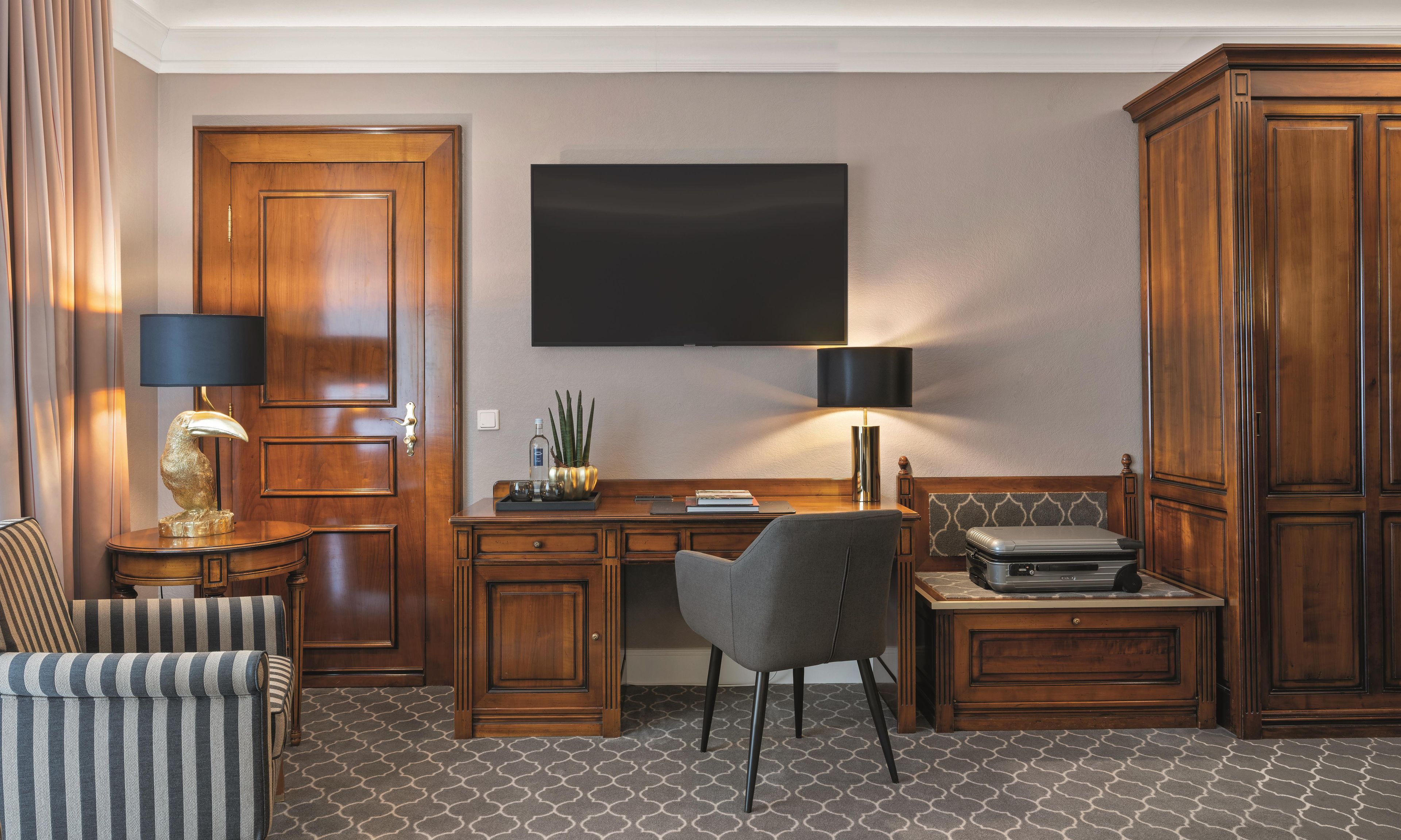 A hotel room with a wooden desk, a grey chair, a television mounted on the wall, and a large wardrobe.