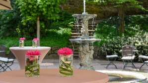 A garden setting with a fountain, two tables with vases of flowers, and outdoor seating. - Parkhotel Engelsburg (Recklinghausen)