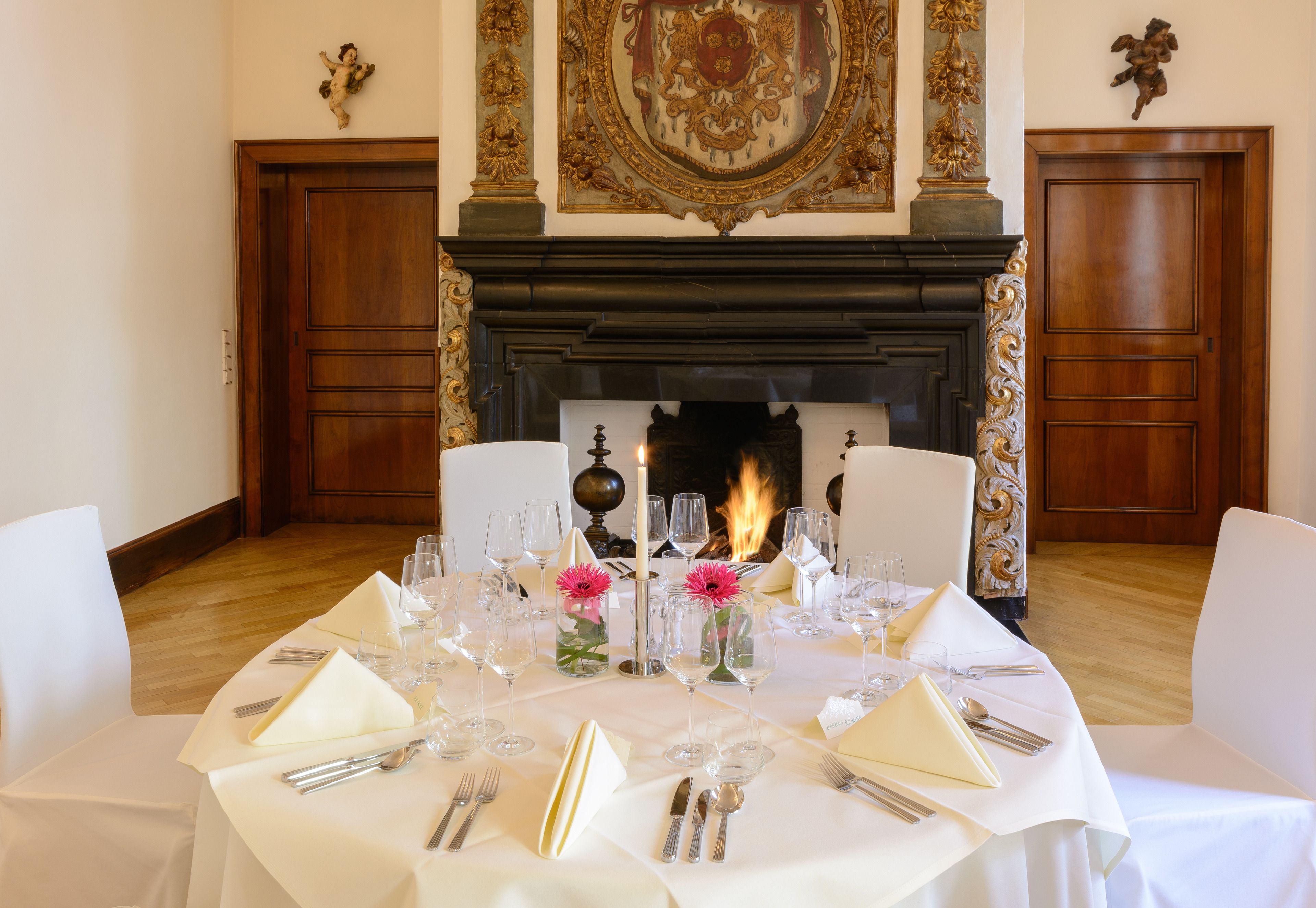 A formal dining table set for a meal with white tablecloths, silverware, and glassware, positioned in front of a fireplace with a decorative mantle and two wooden doors.