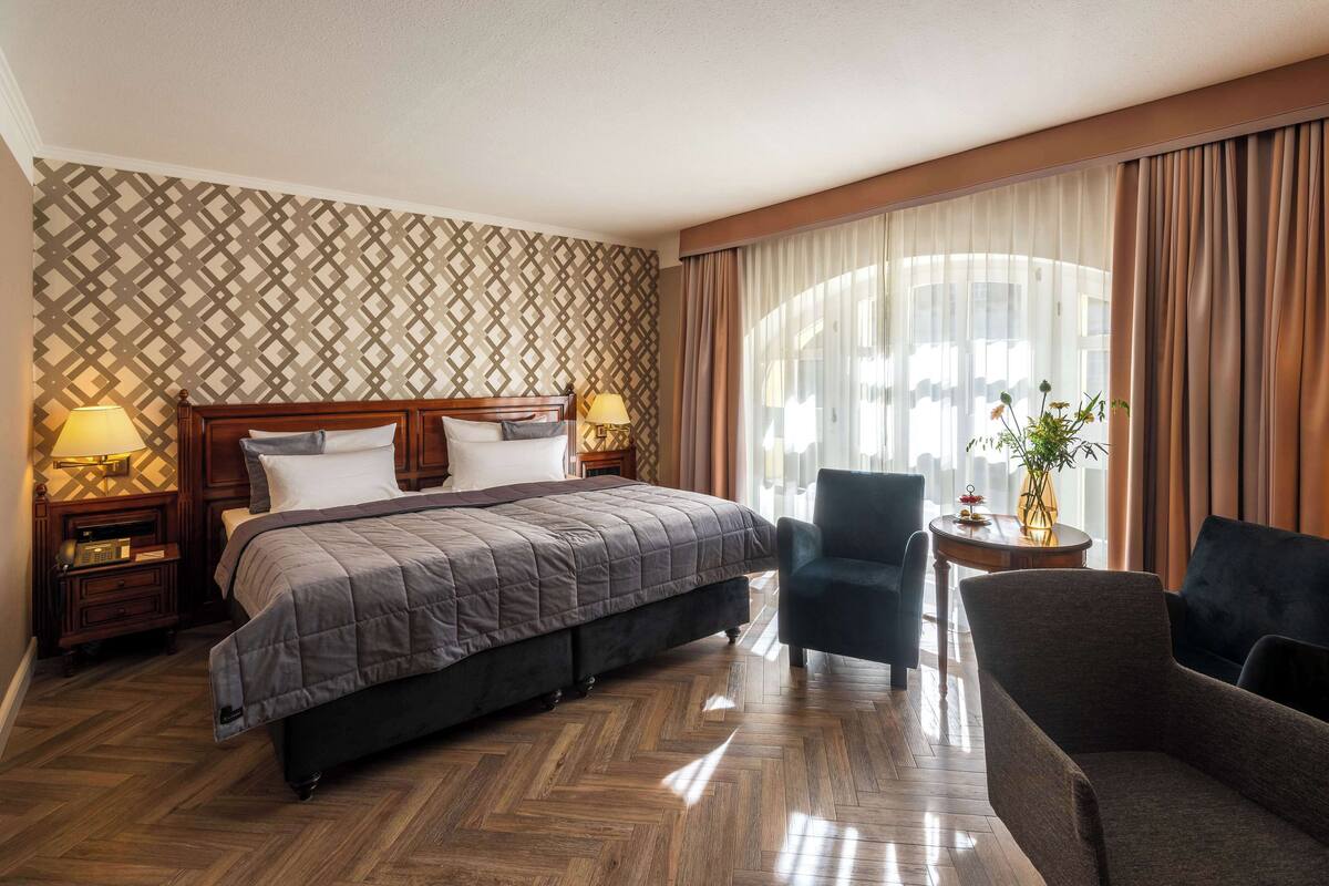 A hotel room with a large bed, two bedside lamps, a chair, a small table, and a window with curtains.