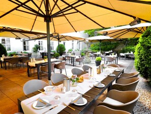 Un área de comedor al aire libre con mesas preparadas para el servicio, sombrillas que dan sombra y una fuente como centro de atención.