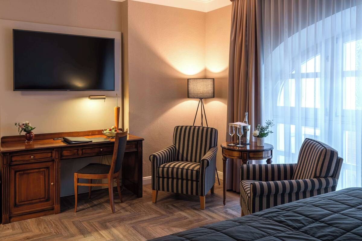 A hotel room with a large flat-screen TV, a desk with a chair, two striped armchairs, a small side table with a lamp, and a bed with a dark quilt.