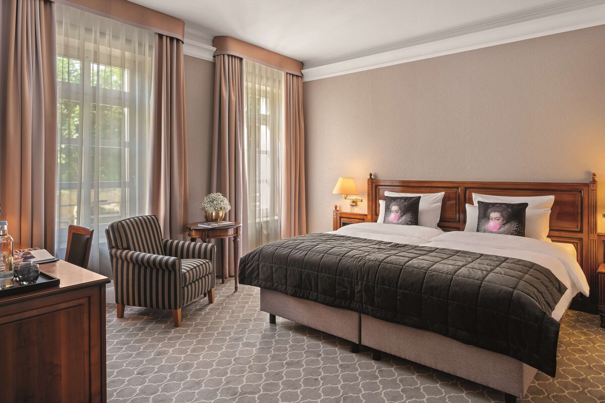 A hotel room with a large bed, a chair, a small table with a lamp, and a view of the outdoors through sheer curtains.