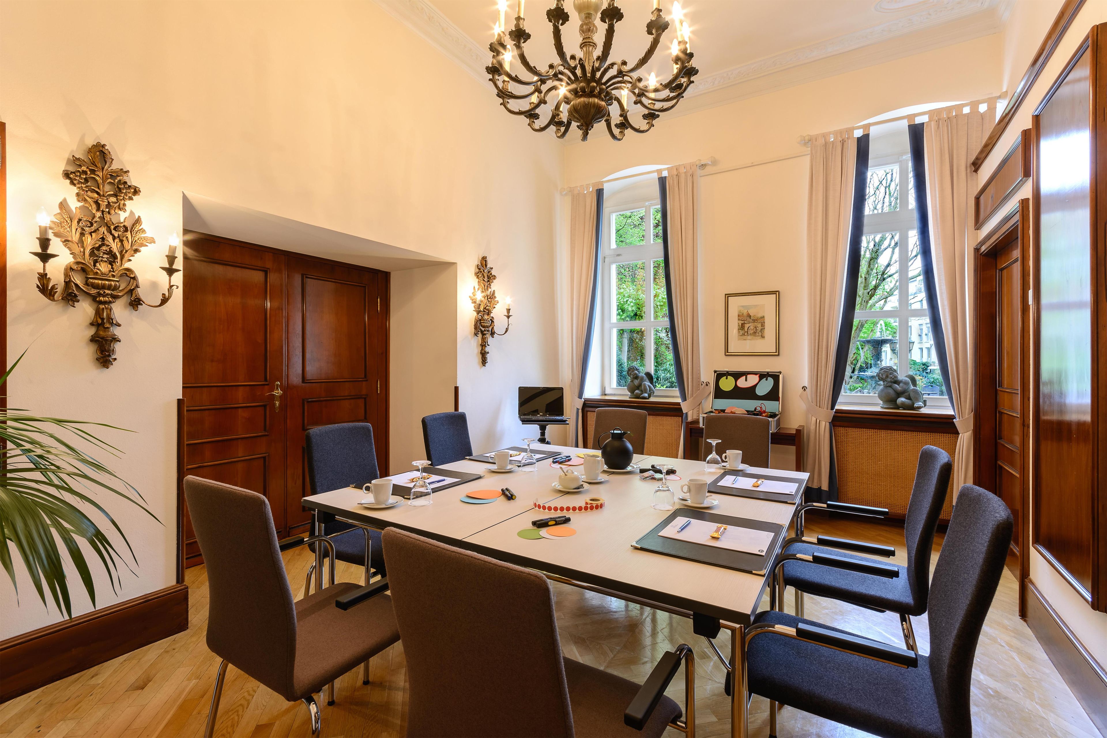 A conference room with a long table, chairs, a chandelier, and large windows with curtains.
