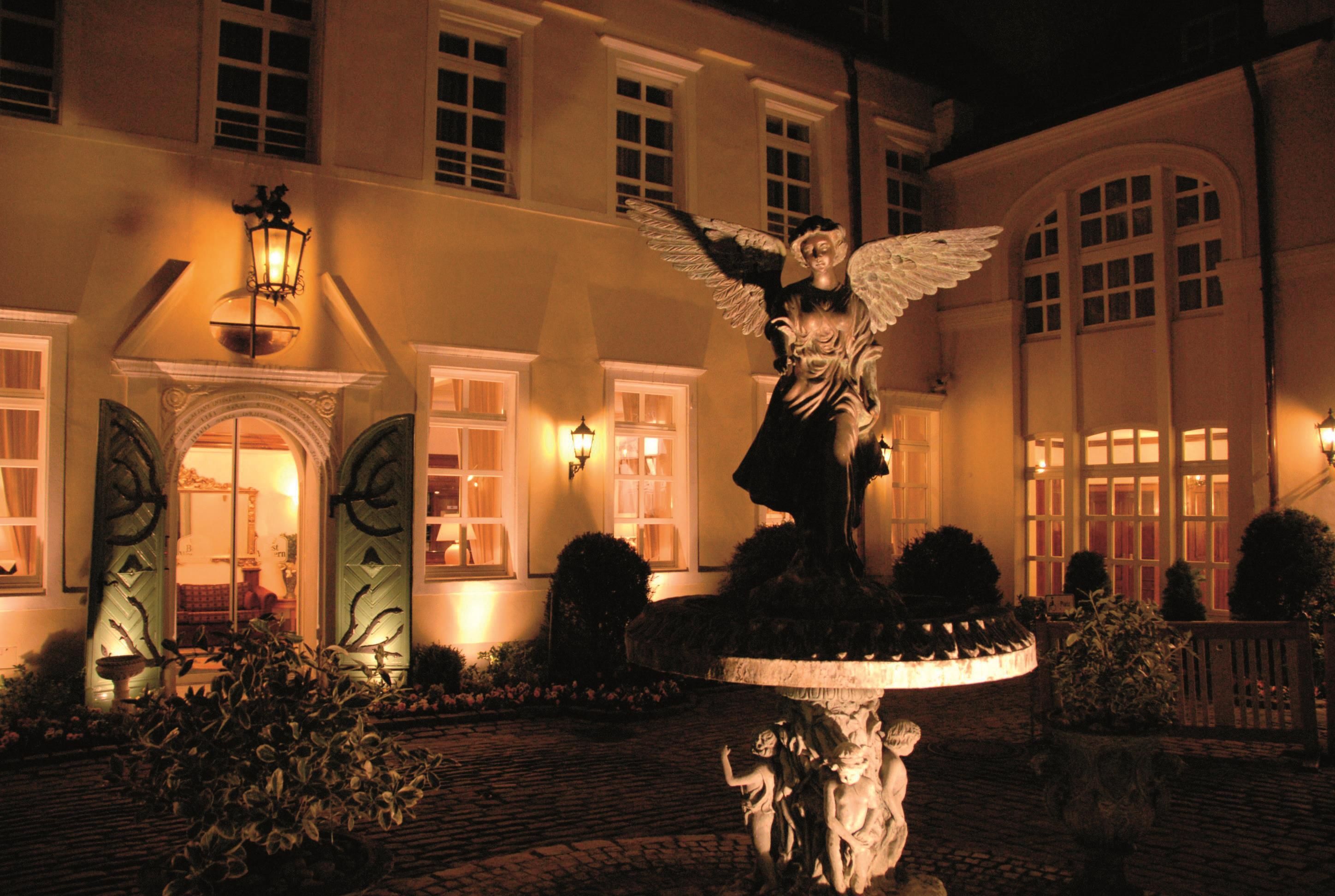Ein Innenhof mit einem Brunnen, der eine Engelsstatue zeigt, umgeben von gepflegten Hecken und einem Gebäude mit zahlreichen Fenstern.