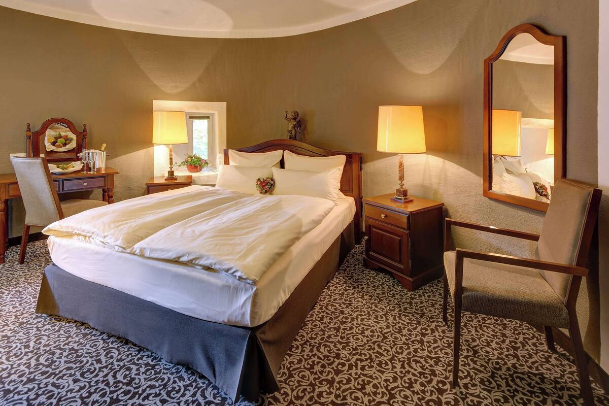 A hotel room with a large bed, a desk with a lamp, a chair, a dresser, and a mirror.