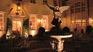 Un patio con una estatua de ángel en una fuente, rodeado de arbustos bien cuidados y un edificio con ventanas grandes y una puerta con un arco decorativo.