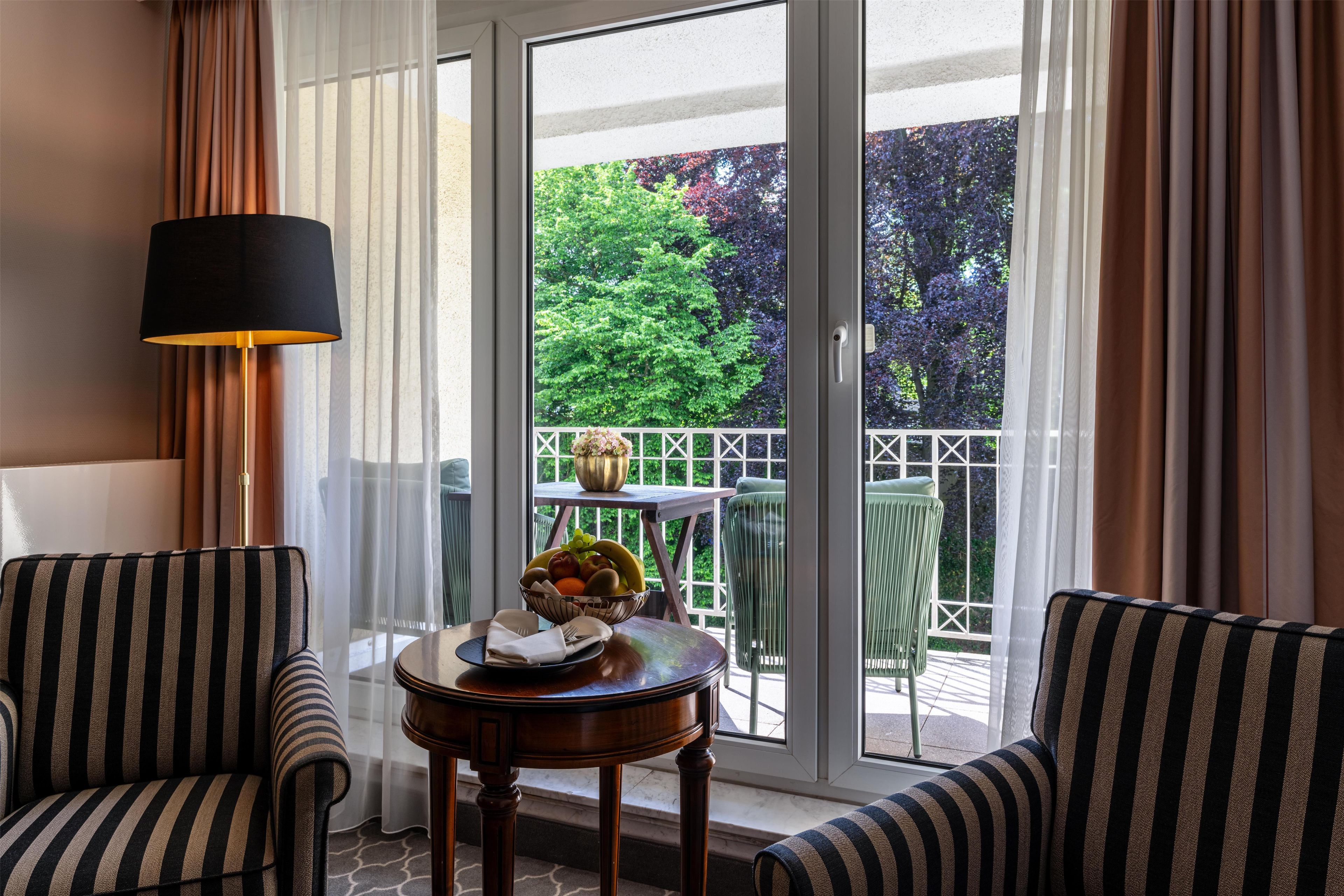 Une pièce avec deux fauteuils à rayures, une petite table ronde sur laquelle se trouve un saladier rempli de fruits, une lampe et une vue sur un jardin, visible à travers une porte vitrée.