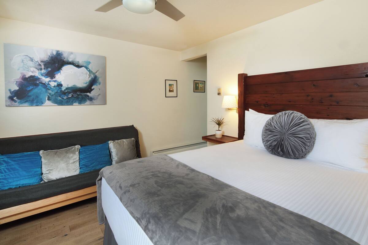 A bedroom with a bed, a sofa, a ceiling fan, and a painting on the wall.