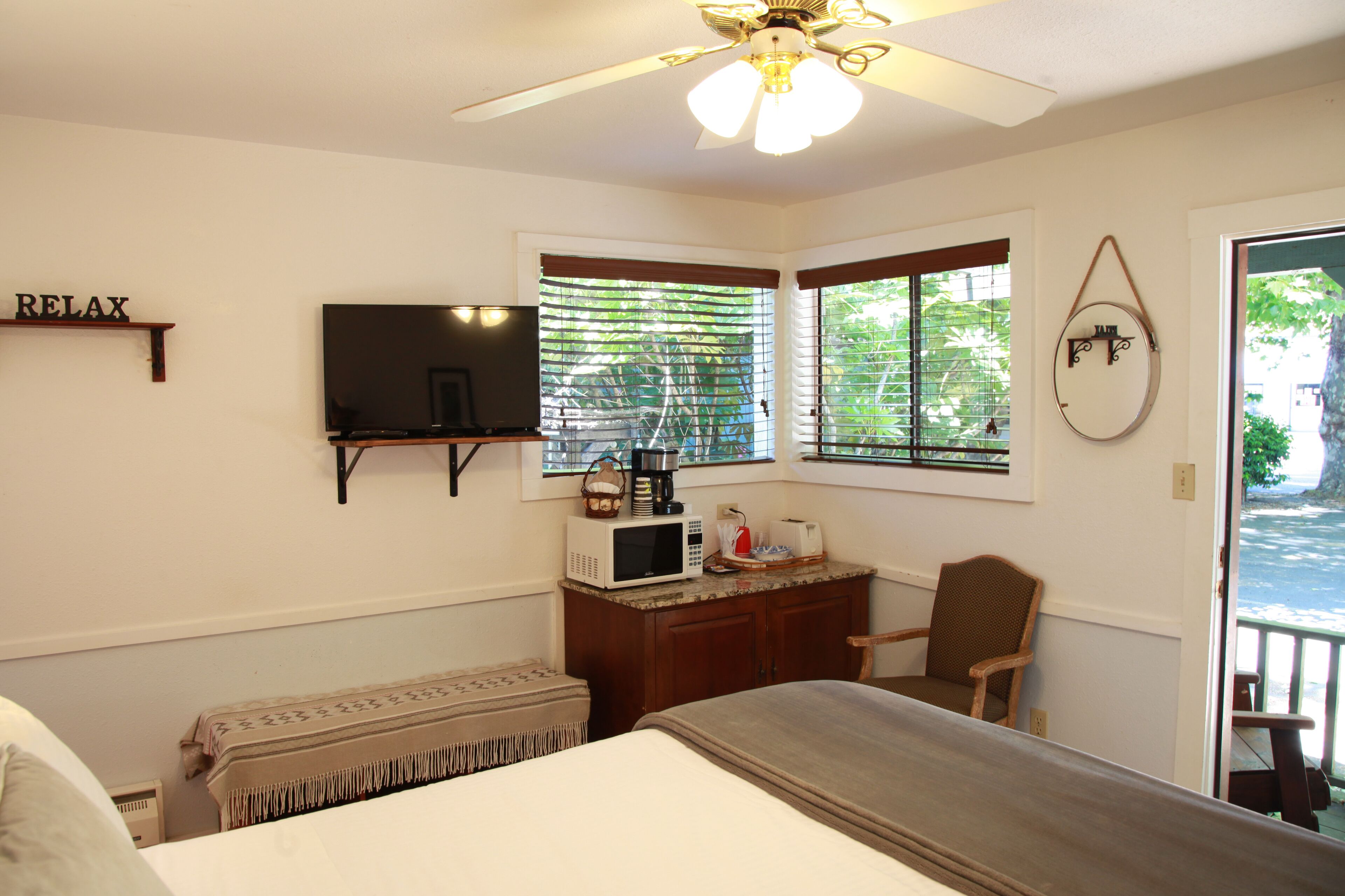 A bedroom with a bed, a desk with a microwave, a chair, a TV, and a window with blinds.