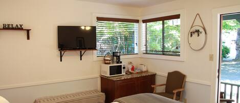 A bedroom with a bed, a desk with a microwave, a chair, a TV, and a window with blinds.