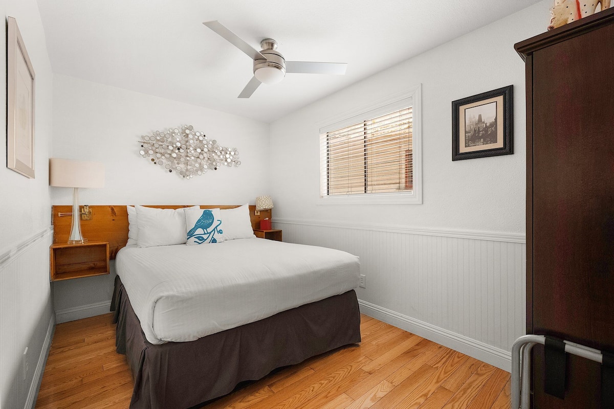 A bedroom with a bed, a ceiling fan, a wall-mounted lamp, a window with blinds, and a framed picture on the wall.